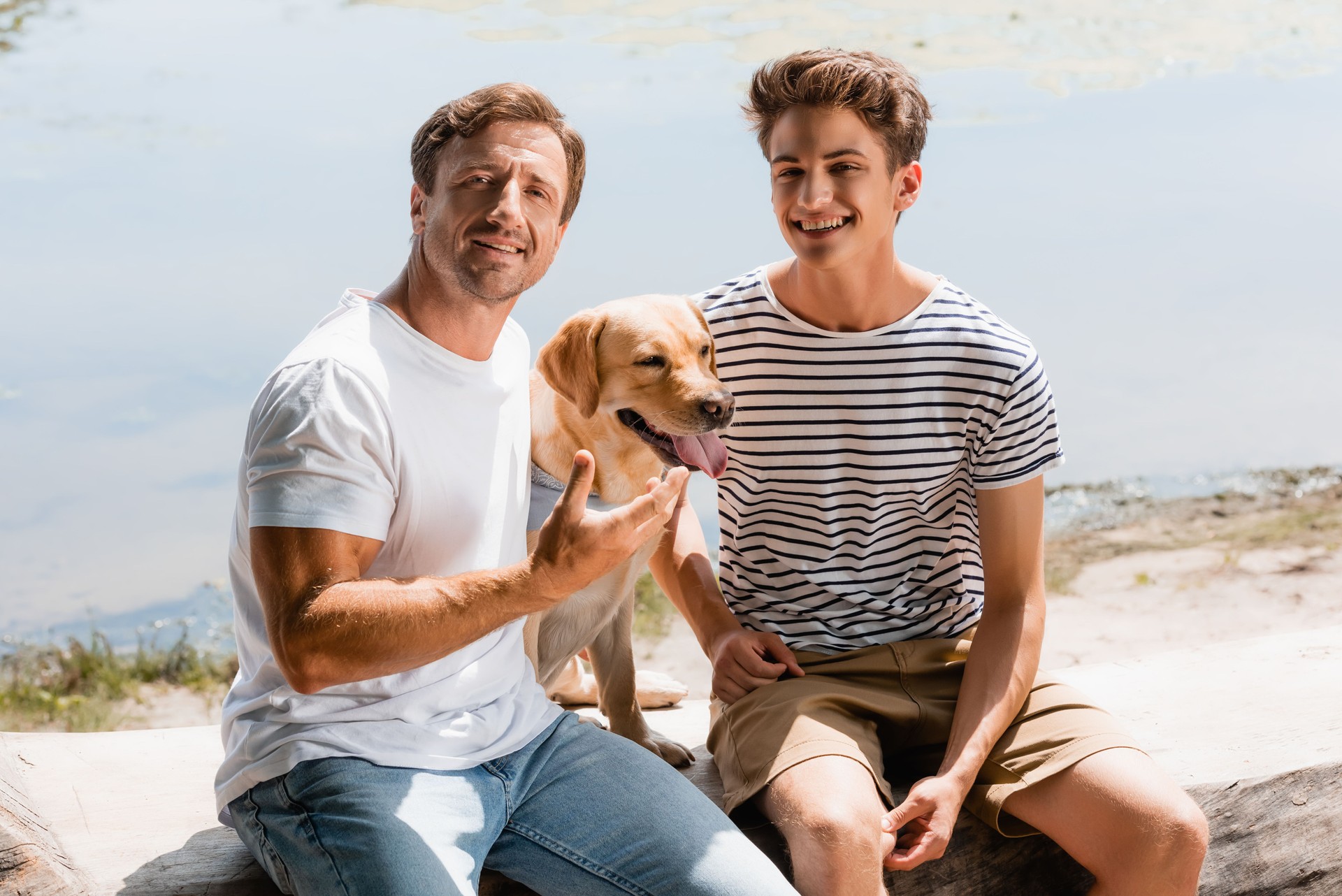 father and son sitting with golden retriever near lake